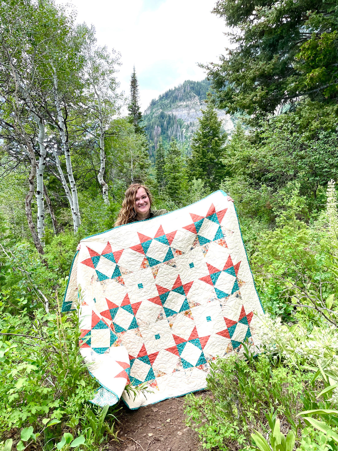 Lilian Star Quilt made of orange and teal fabric on a white background. Maker displaying quilt in a mountainous landscape.
