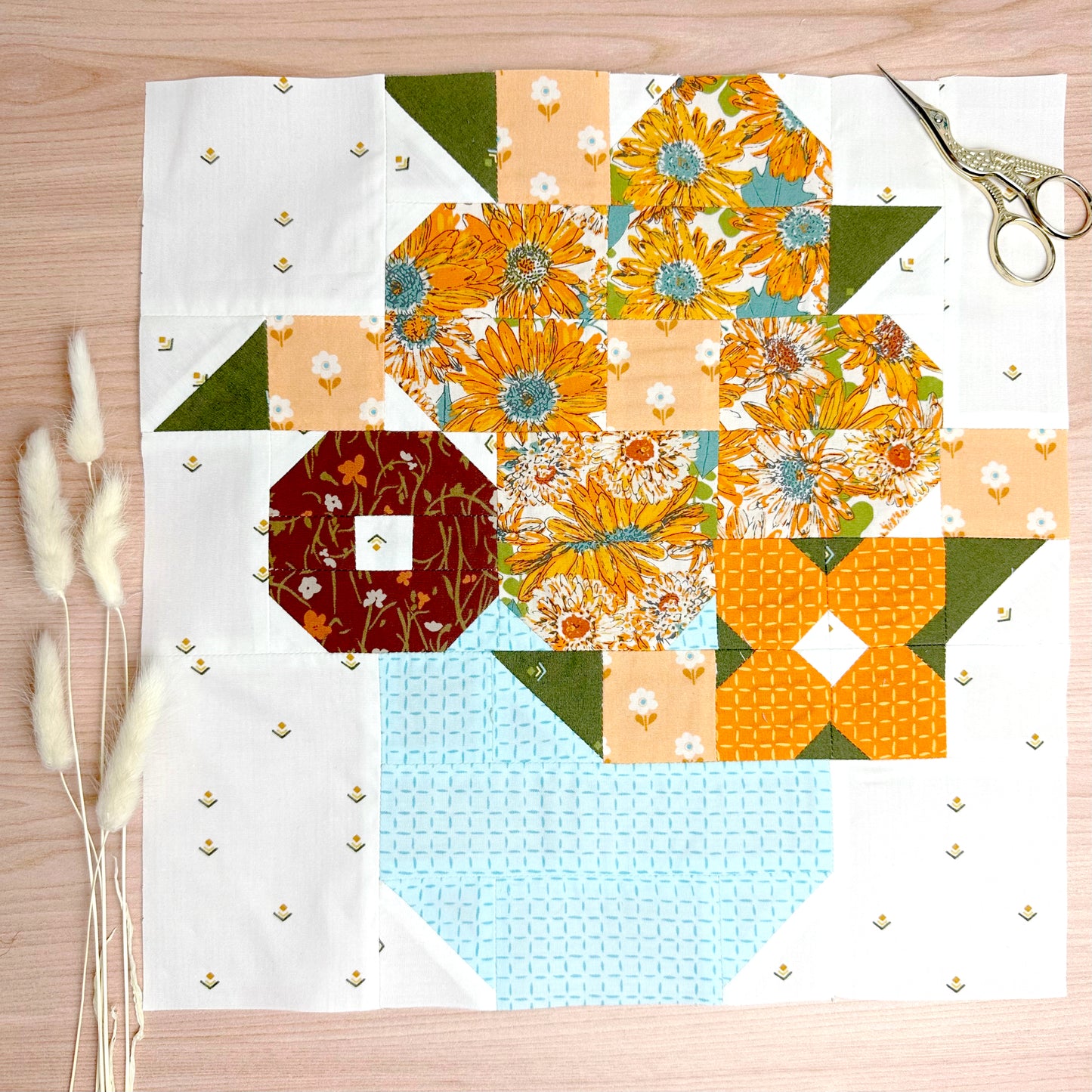 A colorful handmade quilt block featuring floral and geometric patterns, laid flat on a light wood surface with a pair of scissors in the top right and dried bunny tails on the left.