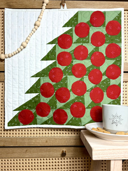 Merry Mini Advent quilt. A small quilt resembling the side of a green patchwork Christmas tree with red circle ornament pockets. Displayed hanging on a wood cabinet. A mug with cookies.