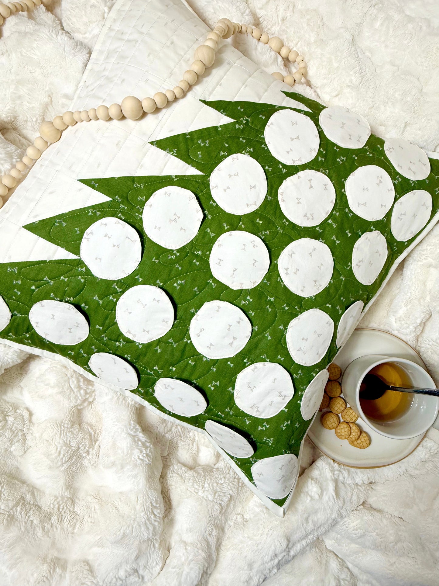 Merry Mini Advent quilt. A small quilt resembling the side of a green patchwork Christmas tree with white circle ornament pockets. Made as a pillow cover. Displayed on a fluffy blanket. A mug with cookies.