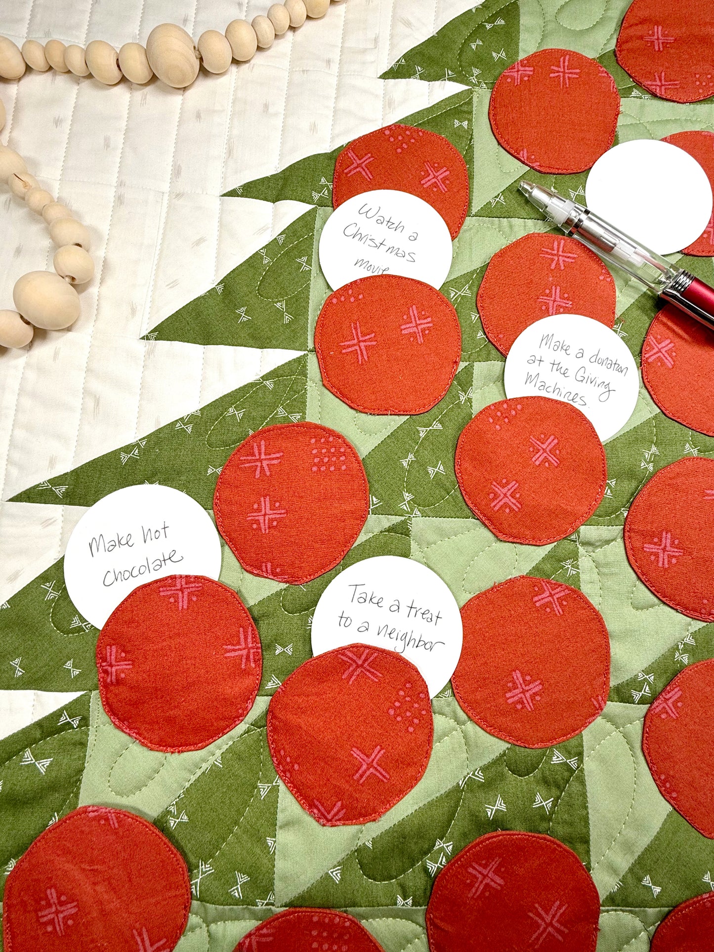Merry Mini Advent quilt. A small quilt resembling the side of a green patchwork Christmas tree with red circle ornament pockets. Displayed flat with paper circles in the pockets with advent activities written on them.