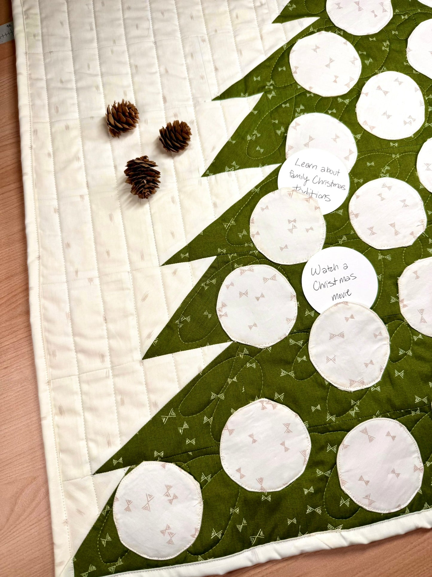 Merry Mini Advent quilt. A small quilt resembling the side of a green patchwork Christmas tree with white circle ornament pockets. Displayed flat with paper circles in the pockets with advent activities written on them.
