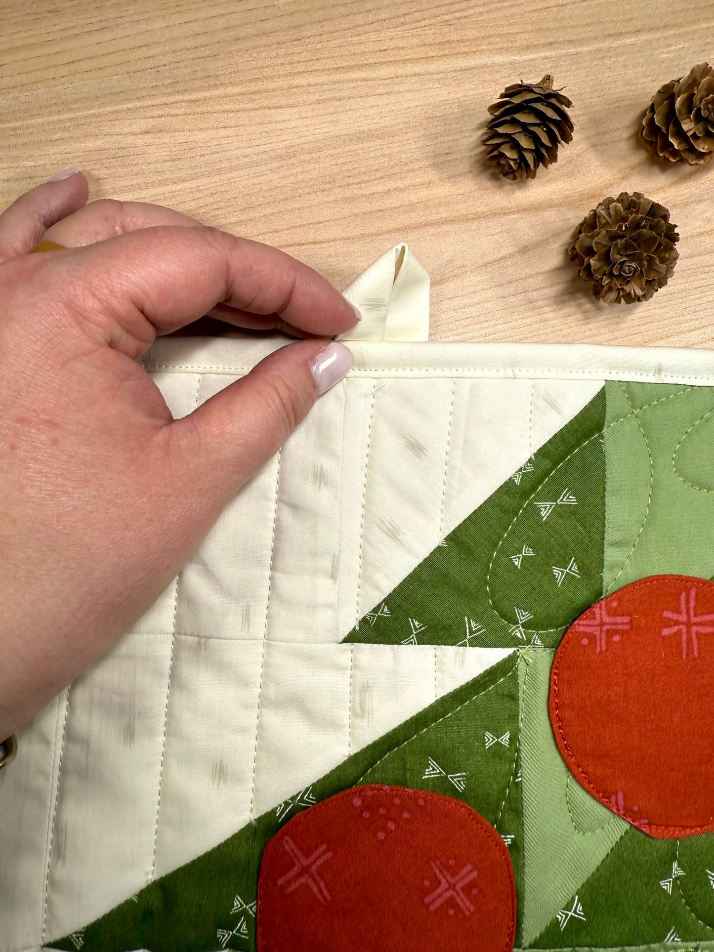 Merry Mini Advent quilt. A small quilt resembling the side of a green patchwork Christmas tree with red circle ornament pockets. Displayed flat, illustrating the sewn-in hook at the top to create a wall hanging. Pinecone props.