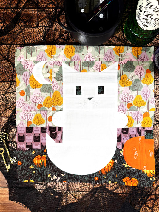 A quilt block featuring a white cat ghost with a pumpkin, moon, and stripe background. Made in orange, purple, green, and black Halloween fabrics. Displayed on a wood background with spooky lace, metal keys, and a glass potion bottle.