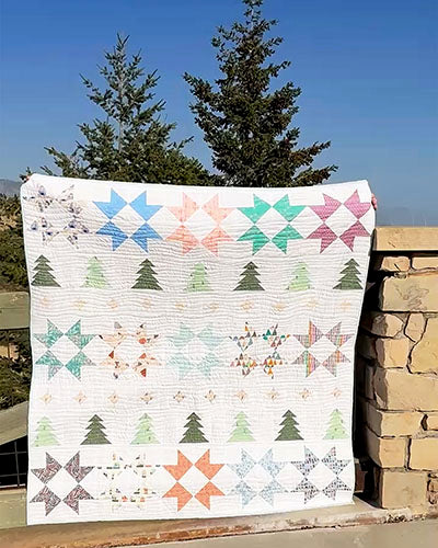 A handmade patchwork quilt with a white background and colorful geometric star and tree patterns, draped outdoors against a stone wall with trees and a blue sky in the background.