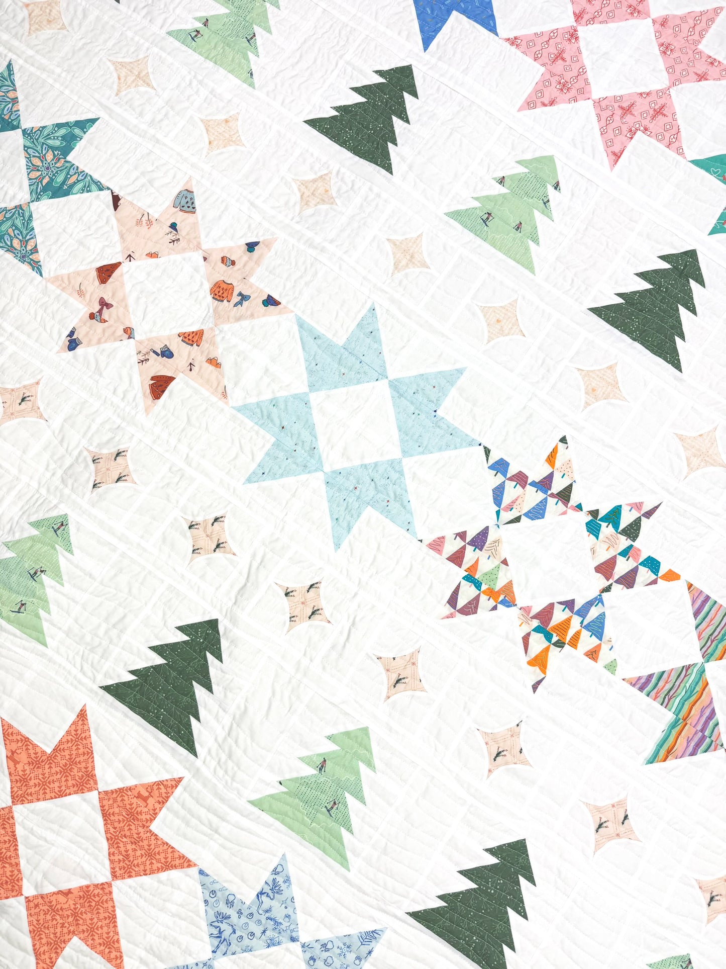 A close-up of a quilt featuring a festive holiday pattern with starbursts, Christmas trees, and snowflakes in various colors on a white background.
