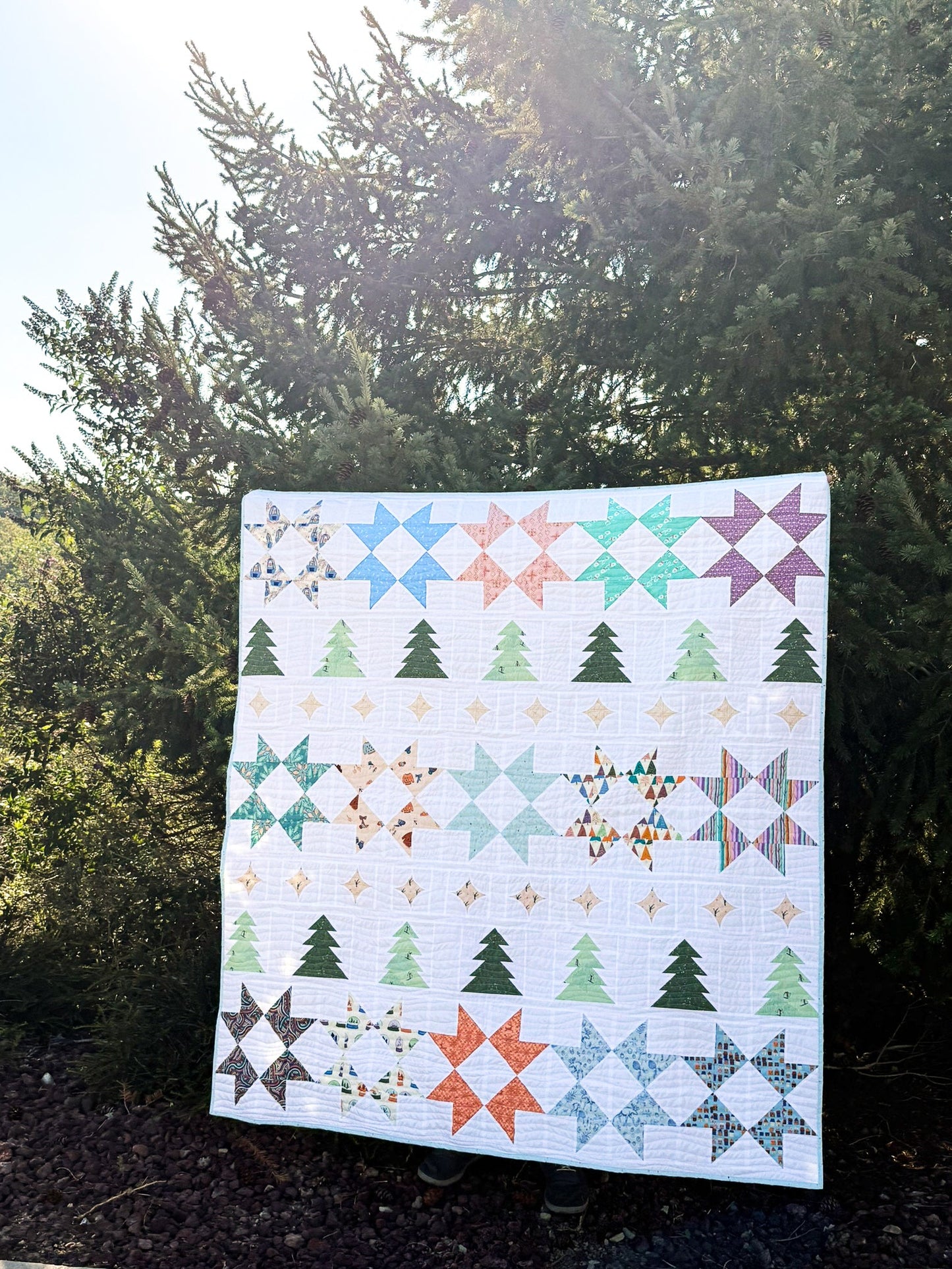 A square quilt with a colorful patchwork pattern of stars, trees, and geometric motifs, held up outdoors against a backdrop of green trees and dark mulch.