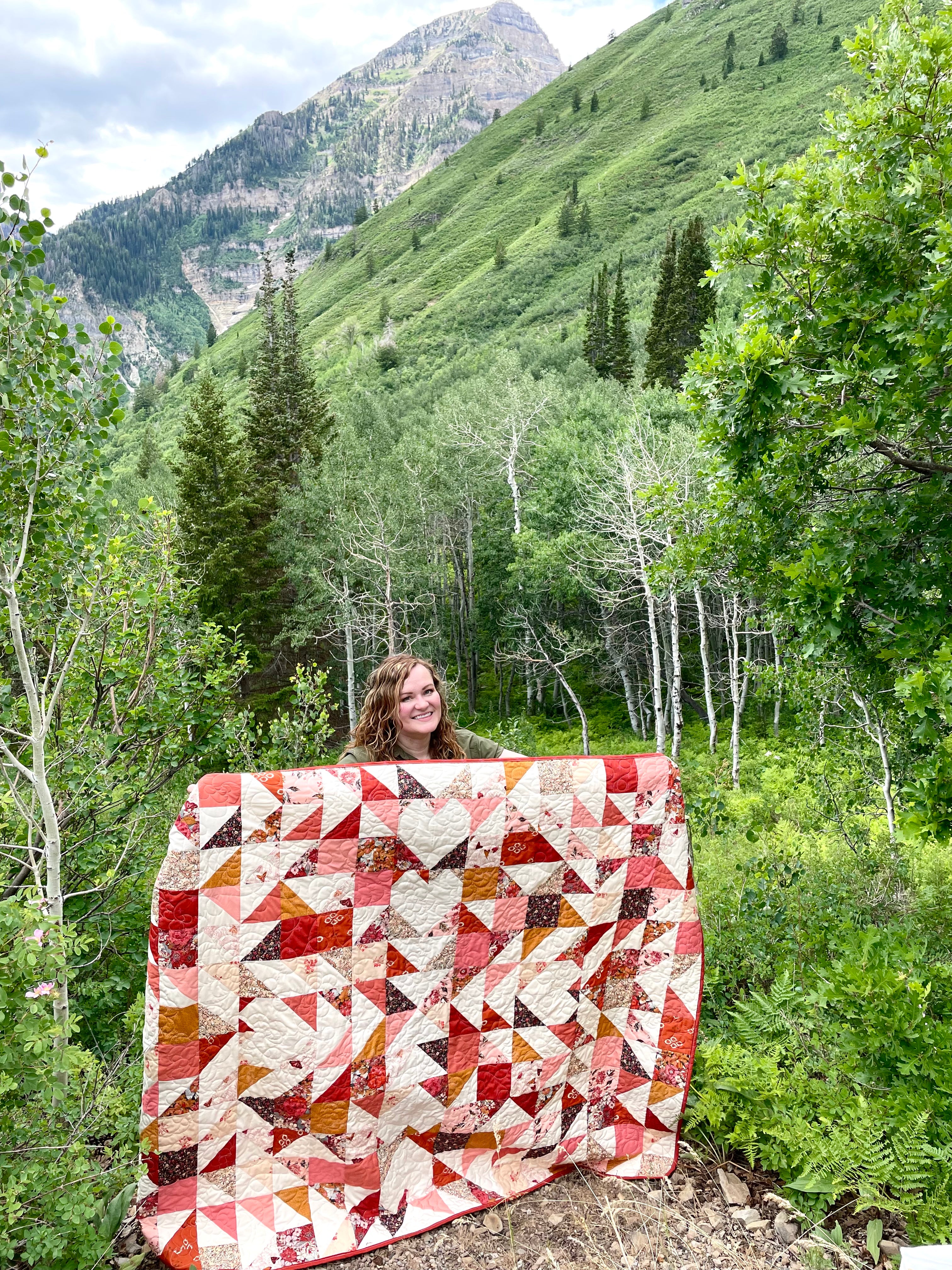Sweet LaNora quilt made of Kismet fabric collection in reds and peach tones. Geometric quilt with hearts and triangles. Displayed by the maker outdoors in a mountainous landscape.