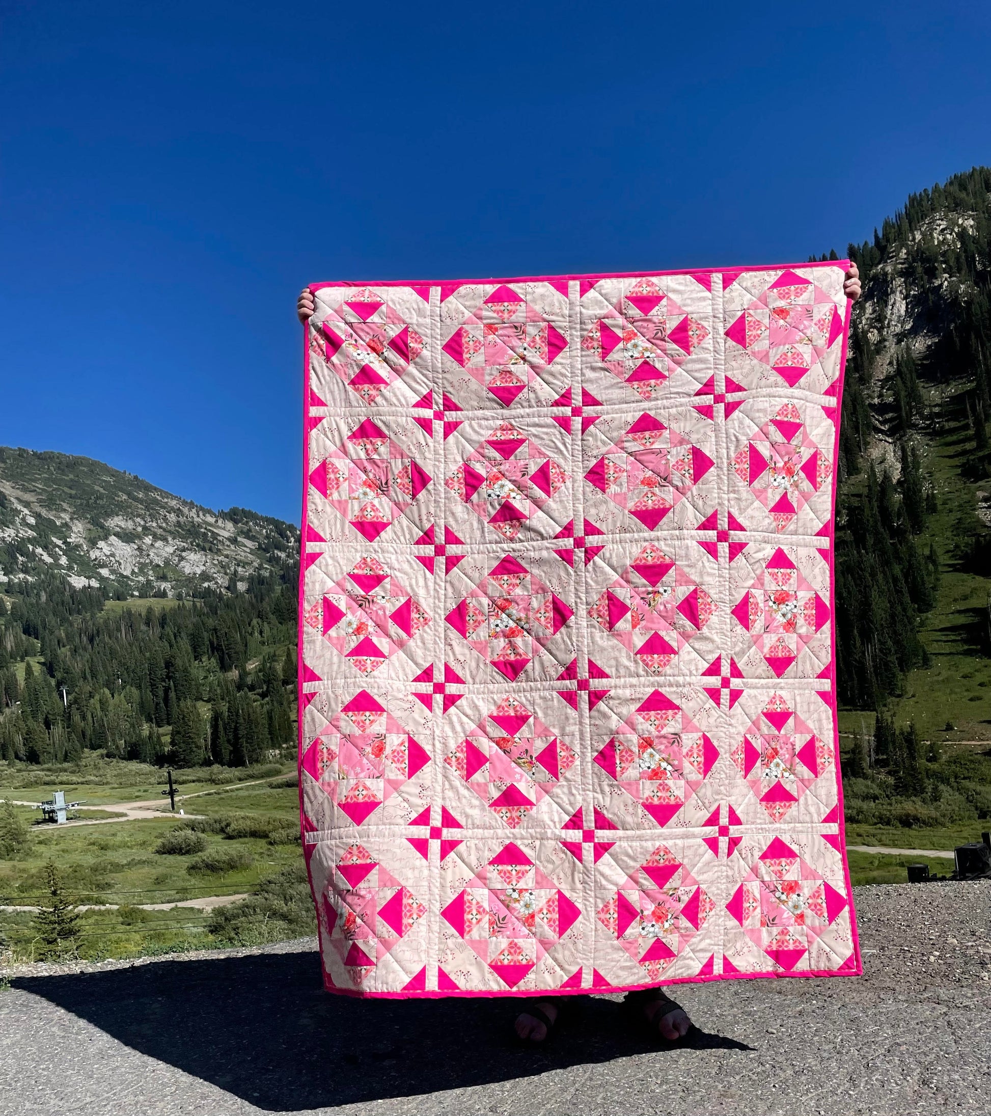 Alta Blooms quilt. A geometric quilt with a small star motif and triangular accents. Bright and medium pinks on a light cream background. Displayed outdoors in a mountain landscape with a blue sky.