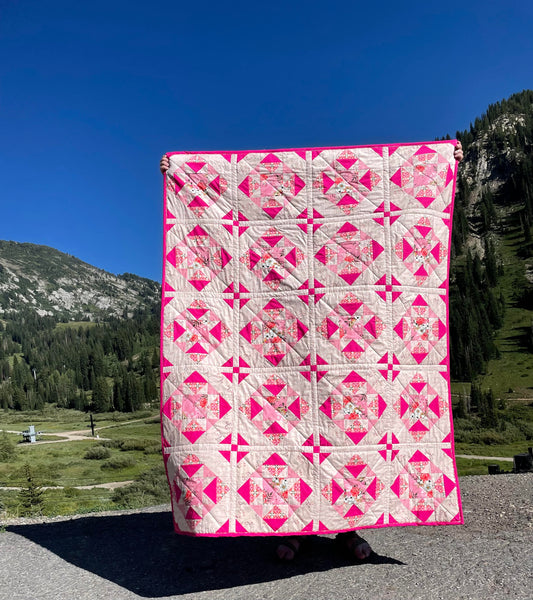 Alta Blooms quilt. A geometric quilt with a small star motif and triangular accents. Bright and medium pinks on a light cream background. Displayed outdoors in a mountain landscape with a blue sky.