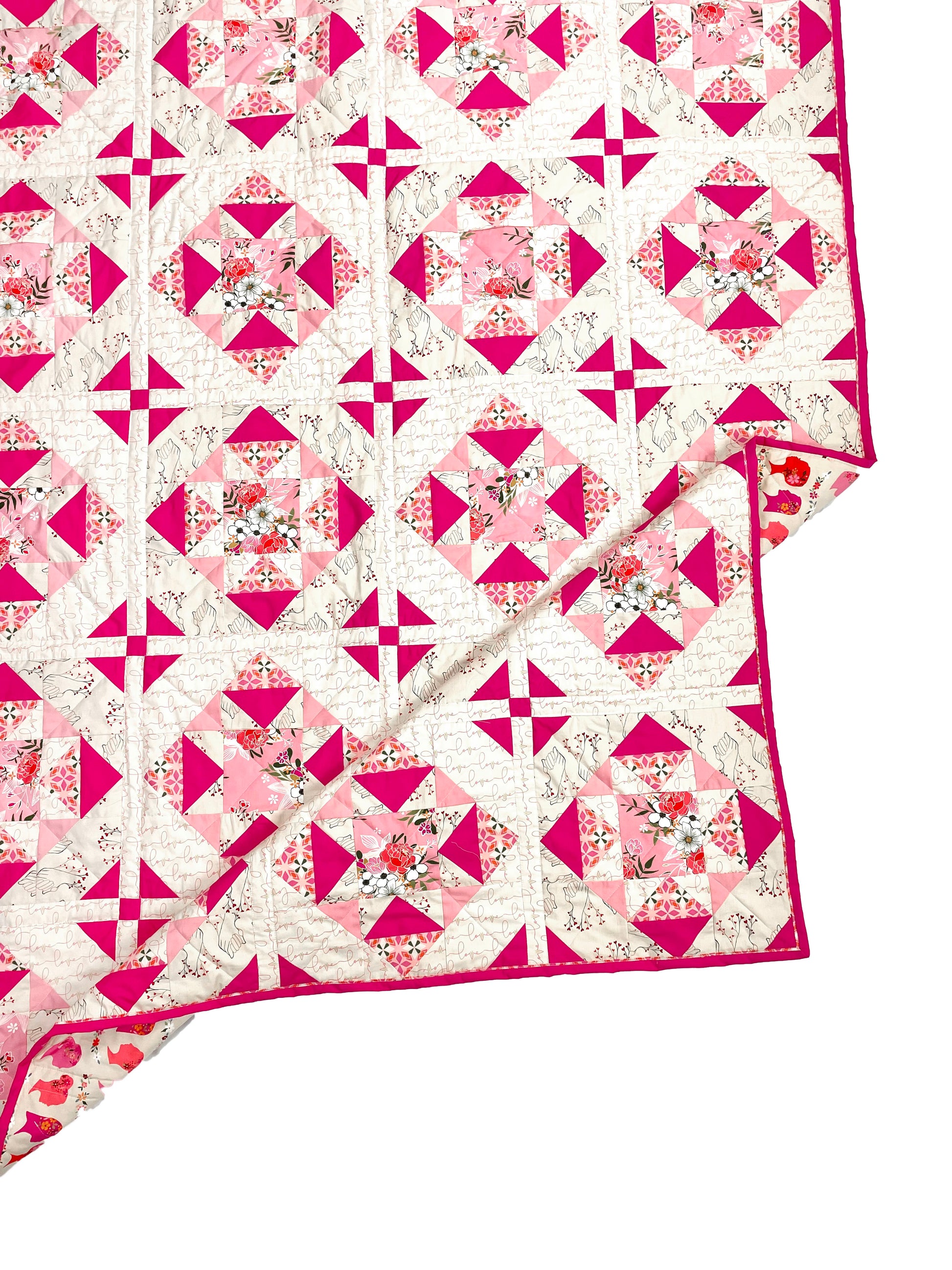 Alta Blooms quilt. A geometric quilt with a small star motif and triangular accents. Bright and medium pinks on a light cream background. Displayed flat on a white surface with the corner folded.