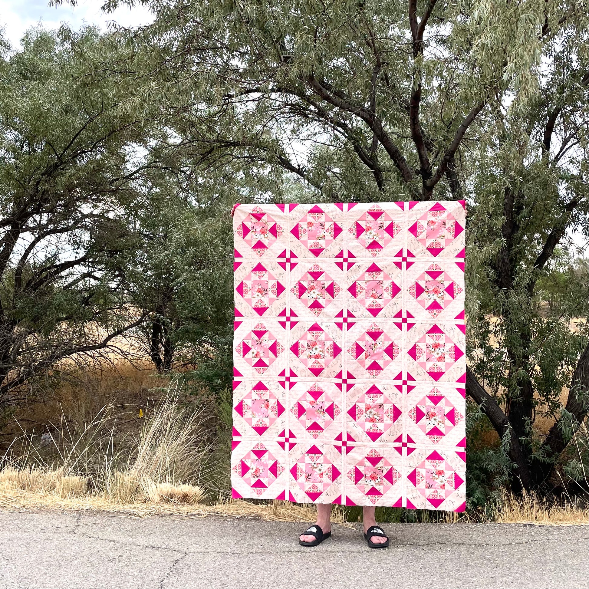 Alta Blooms quilt. A geometric quilt with a small star motif and triangular accents. Bright and medium pinks on a light cream background. Displayed outdoors by a large tree with dark branches.