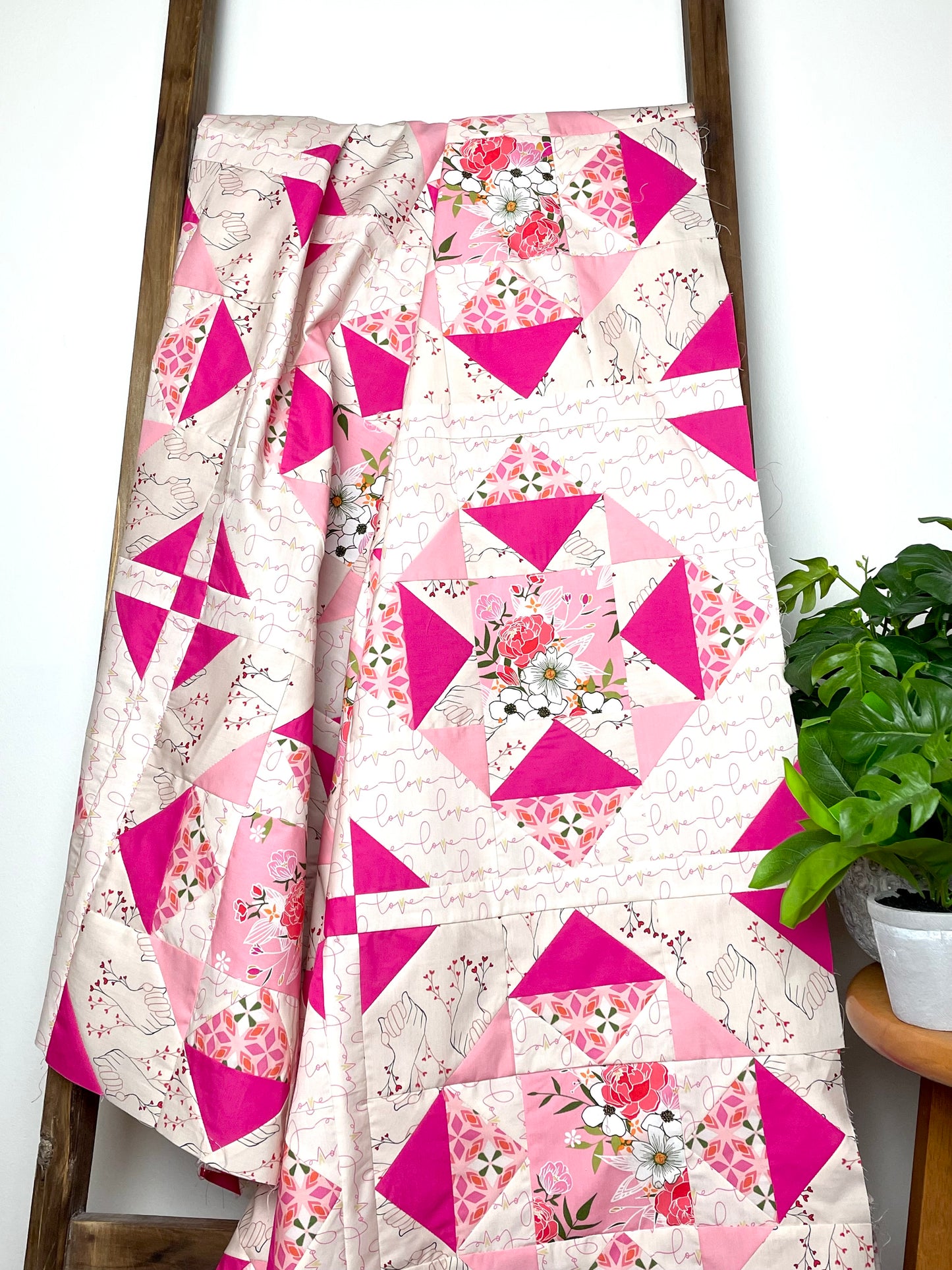 Alta Blooms quilt. A geometric quilt with a small star motif and triangular accents. Bright and medium pinks on a light cream background. Displayed on a brown ladder next to a small, green plant.
