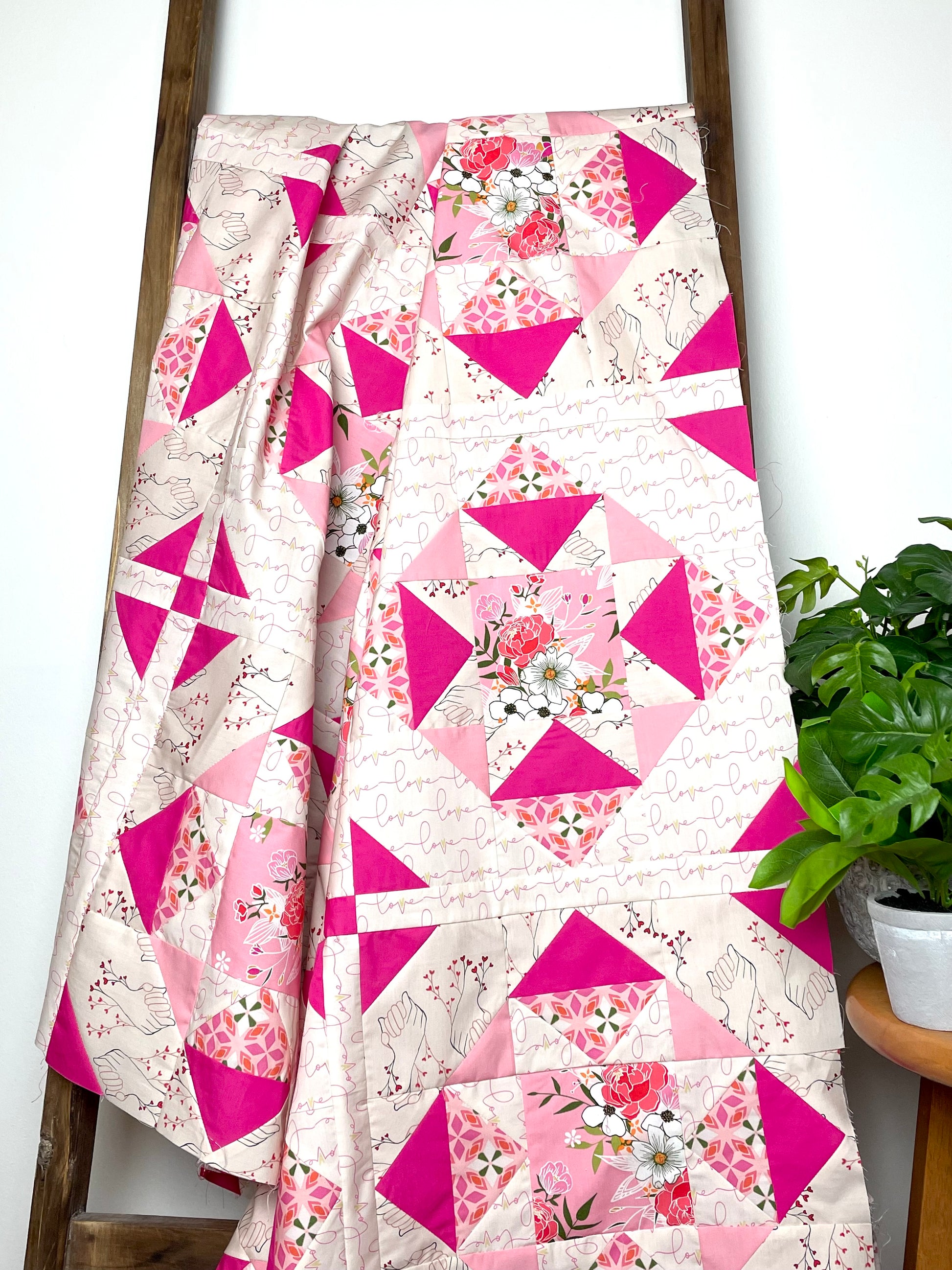 Alta Blooms quilt. A geometric quilt with a small star motif and triangular accents. Bright and medium pinks on a light cream background. Displayed on a brown ladder next to a small, green plant.