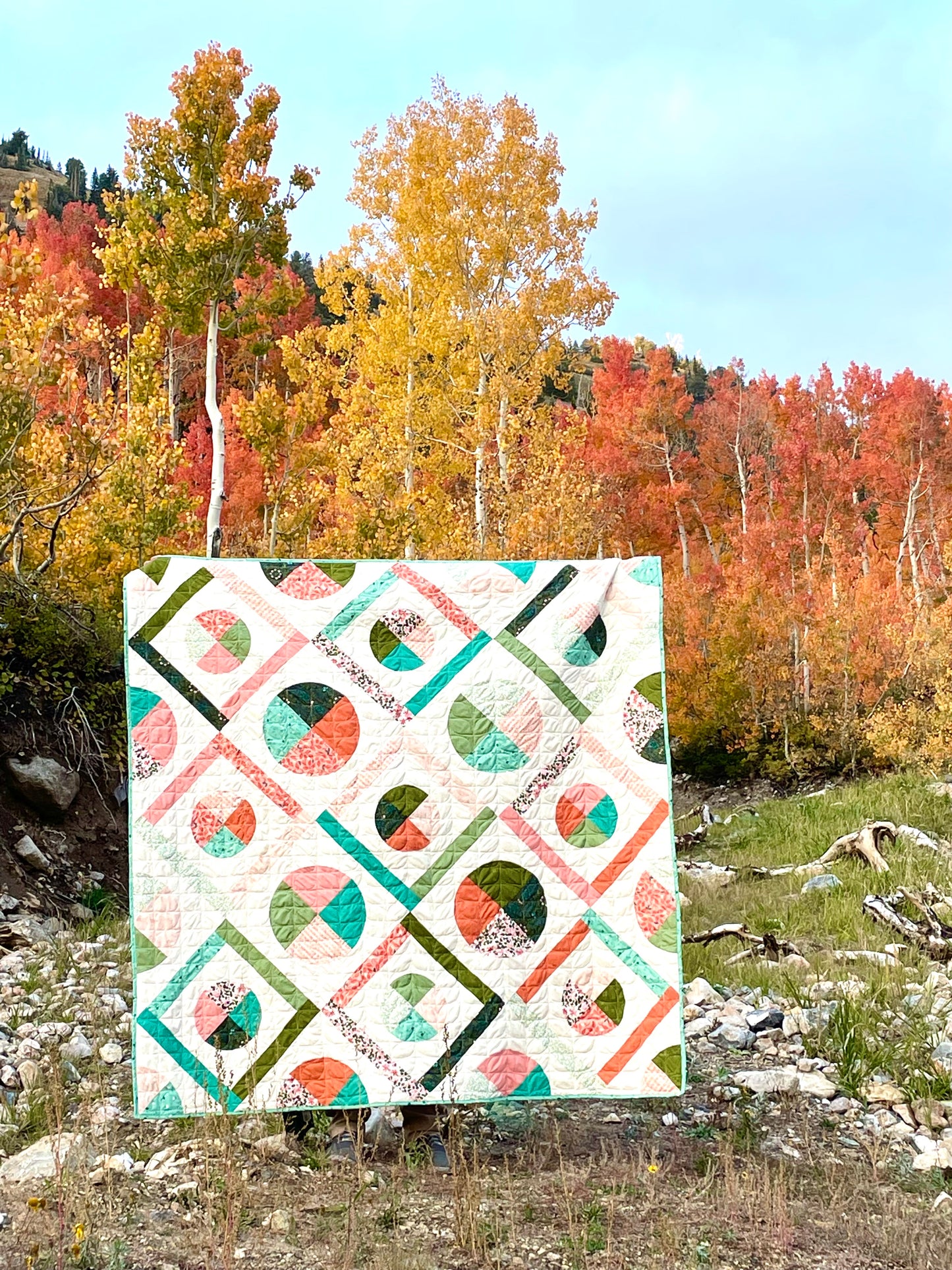 Dora May quilt. A geometric quilt with quarter circles and straight fabric strips in a diamond shape. Made of teal, olive, pink, and orange floral fabrics on a light background. Displayed outdoors by trees in autumn.