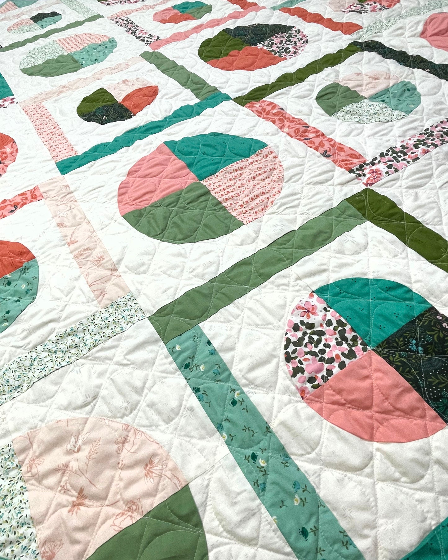 Dora May quilt. A geometric quilt with quarter circles and straight fabric strips in a diamond shape. Made of teal, olive, pink, and orange floral fabrics on a light background. Displayed flat. A close-up.