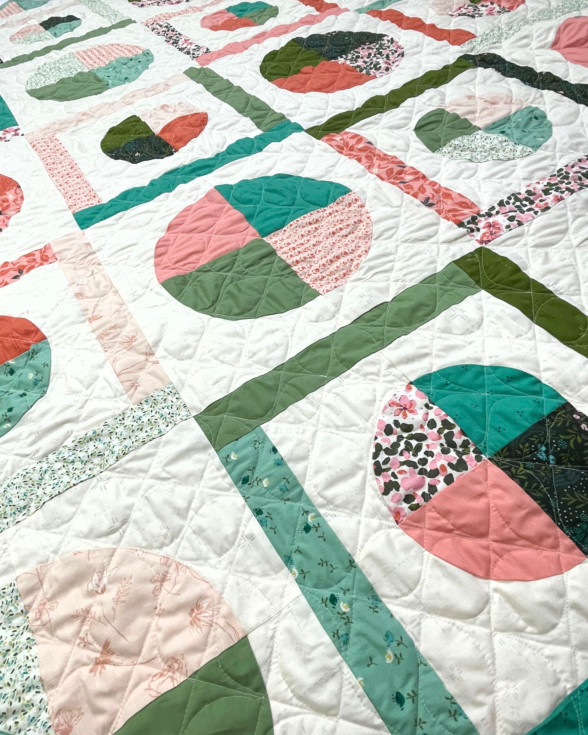 Dora May quilt. A geometric quilt with quarter circles and straight fabric strips in a diamond shape. Made of teal, olive, pink, and orange floral fabrics on a light background. Displayed flat. A close-up.