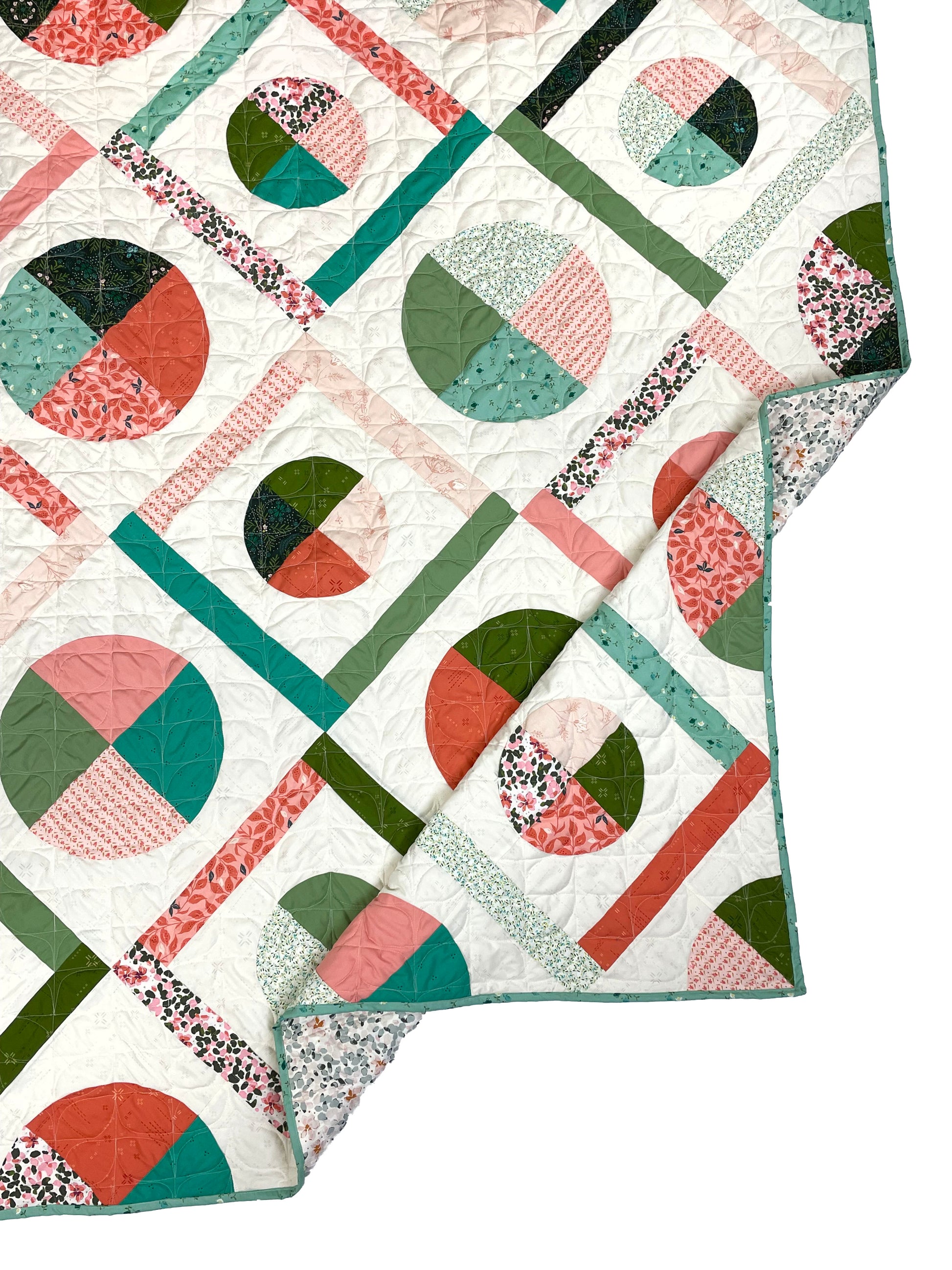 Dora May quilt. A geometric quilt with quarter circles and straight fabric strips in a diamond shape. Made of teal, olive, pink, and orange floral fabrics on a light background. Displayed flat on a white surface with the corner folded.