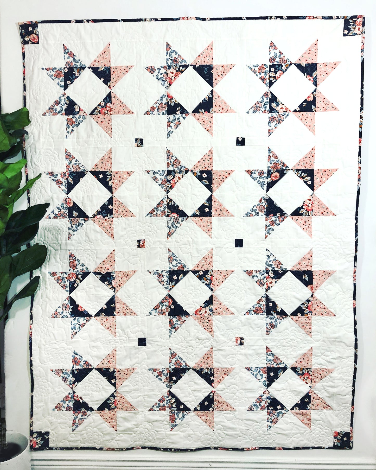Lilian Star quilt. A peach and navy star quilt with a white background. Displayed on a wall with a green ficus plant to the side.