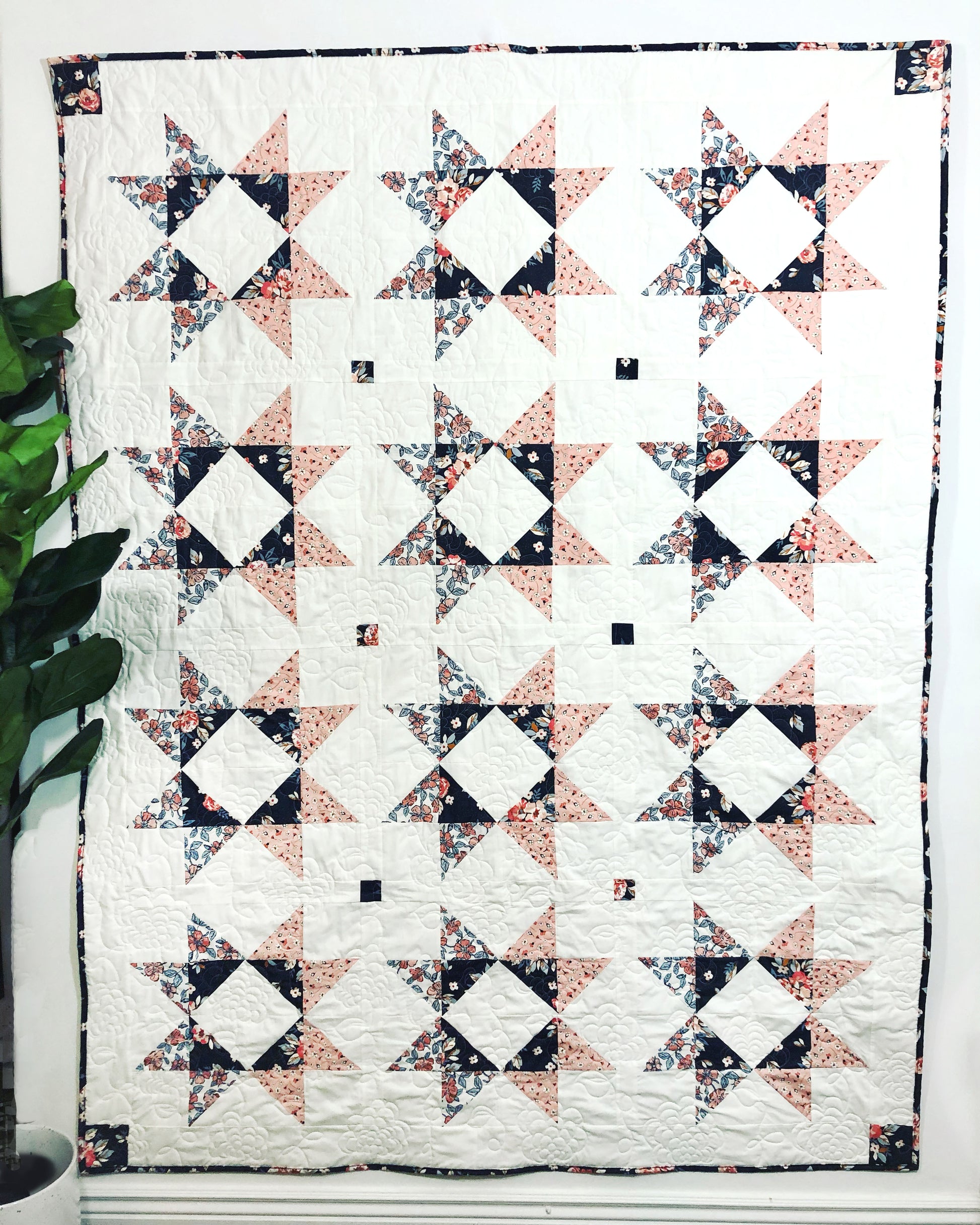 Lilian Star quilt. A peach and navy star quilt with a white background. Displayed on a wall with a green ficus plant to the side.