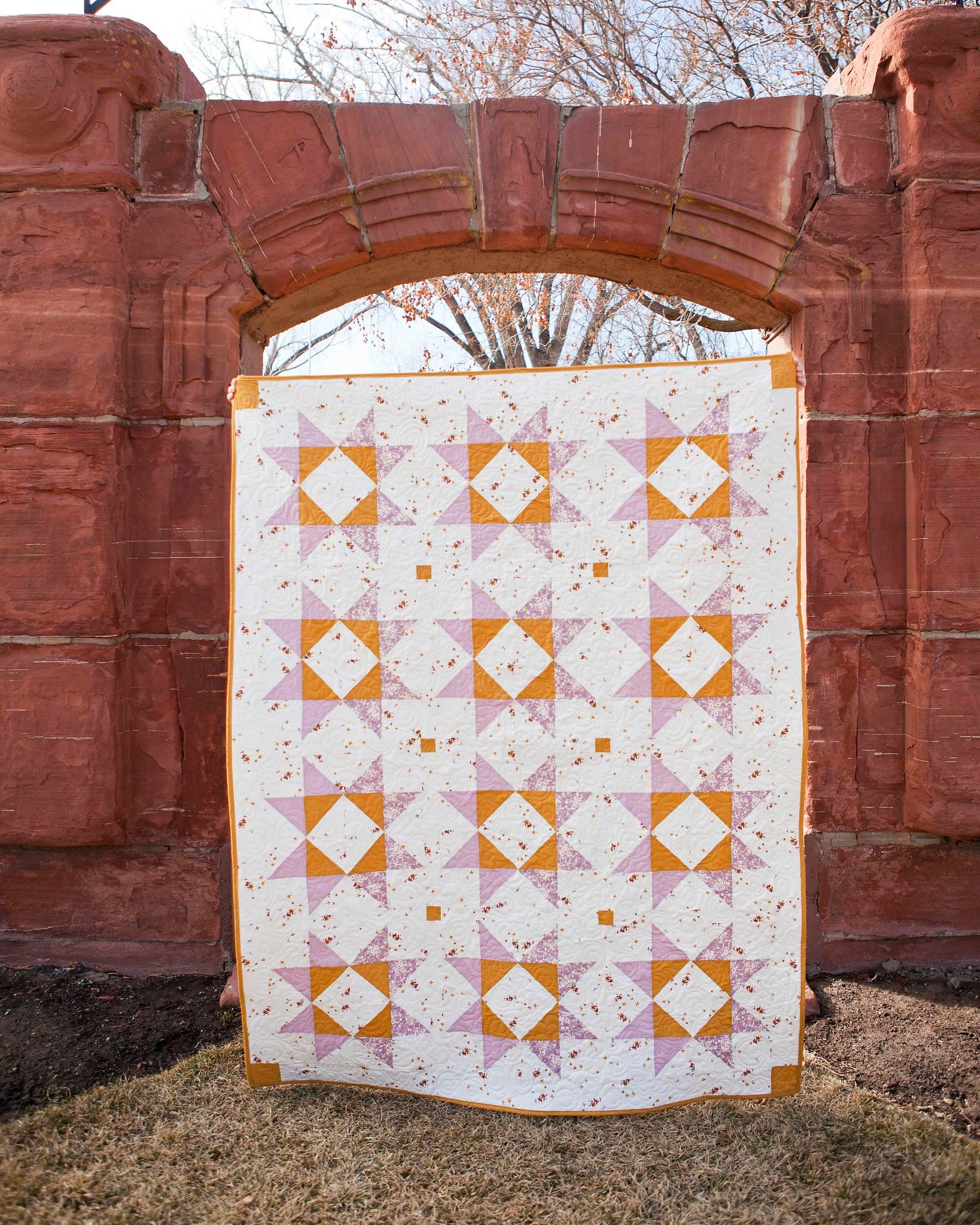 Lilian Star quilt. A geometric star quilt made with yellow and light purple fabrics on a white background. Displayed outdoors under an orange stone arch.