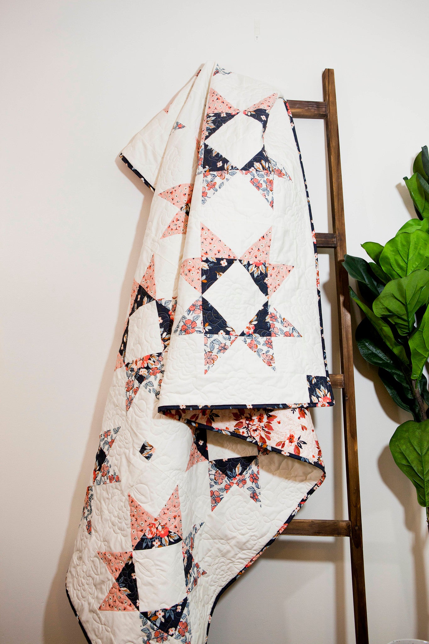 Lilian Star quilt. A geometric star quilt made with peach and navy fabric with a white background. Displayed indoors against a white wall on a brown blanket ladder. A ficus plant displayed on the side.