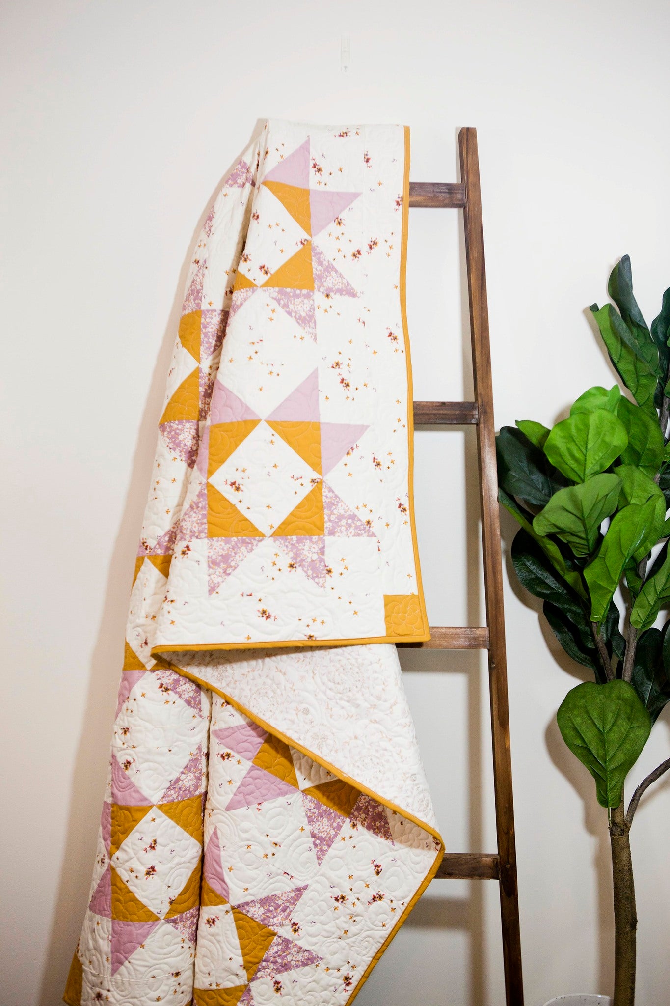 Lilian Star quilt. A geometric star quilt made with yellow and light purple fabrics on a white background. Displayed indoors against a white wall on a brown blanket ladder. A ficus plant displayed on the side.
