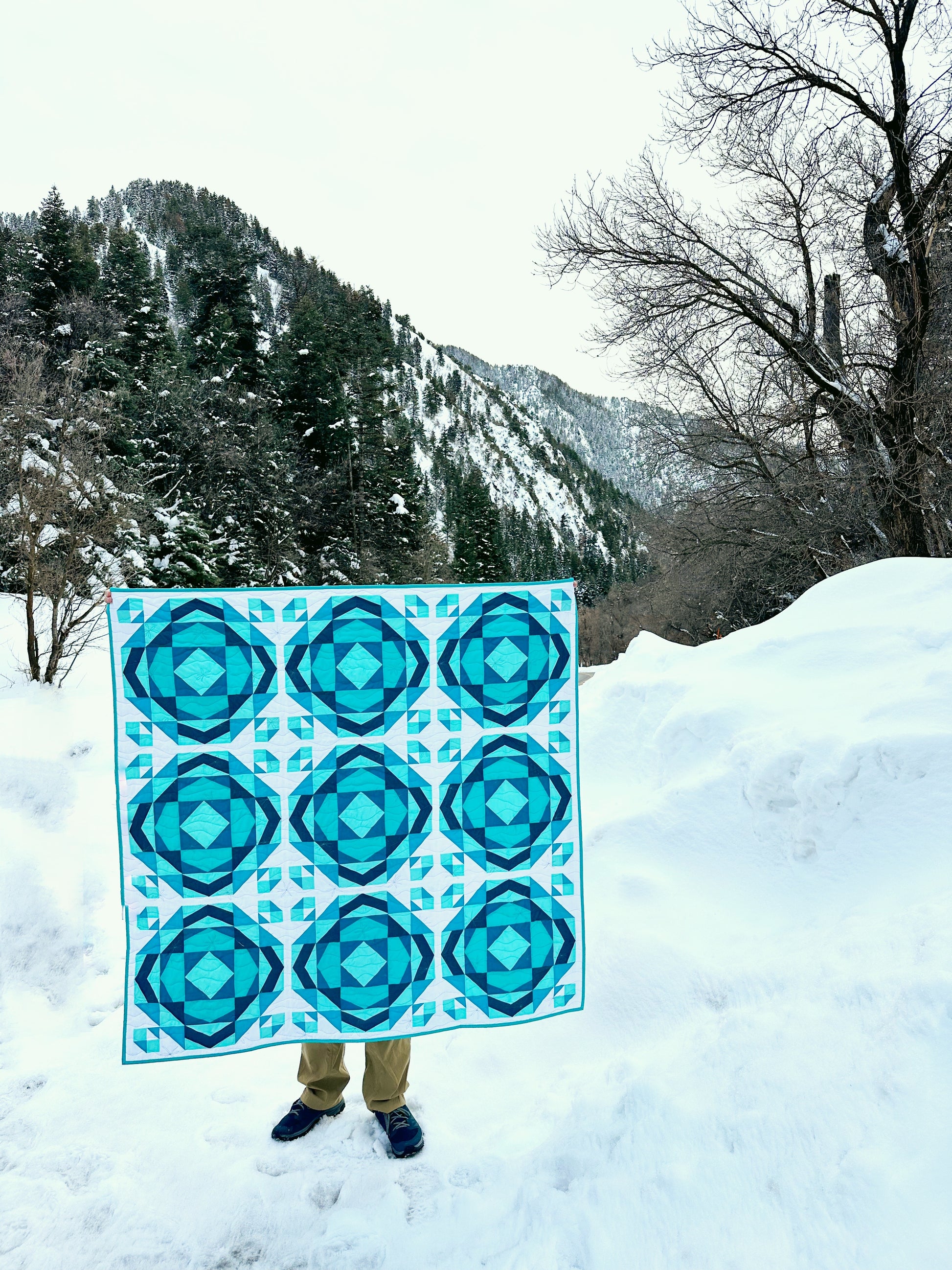 Millcreek quilt. A geometric quilt with a repeating motif that mimics water ripples. Made in varied shades of teal blue with a white background. Displayed outdoors in a snowy mountainous landscape.