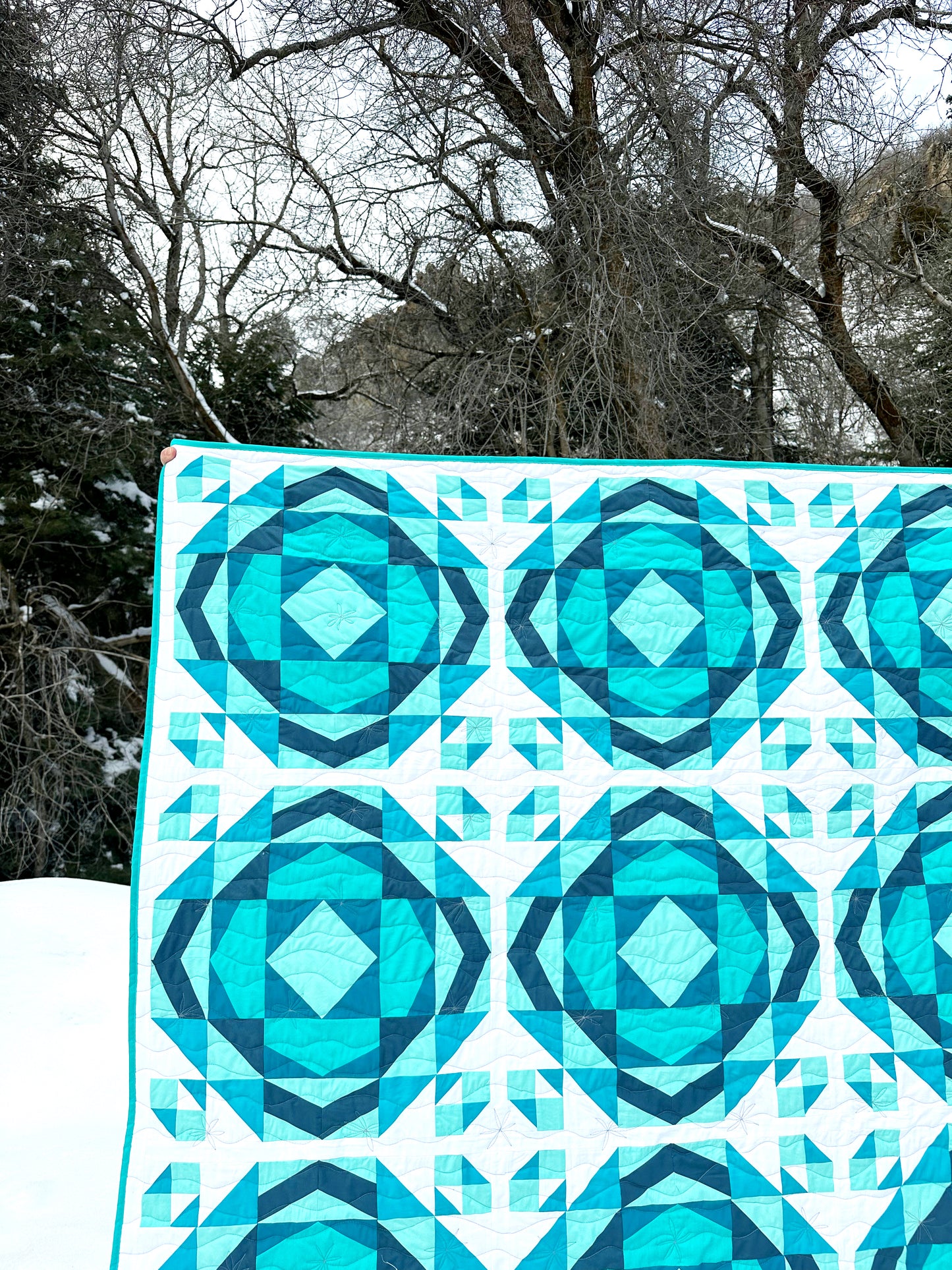 Millcreek quilt. A geometric quilt with a repeating motif that mimics water ripples. Made in varied shades of teal blue with a white background. Displayed outdoors in a snowy mountainous landscape in front of trees.