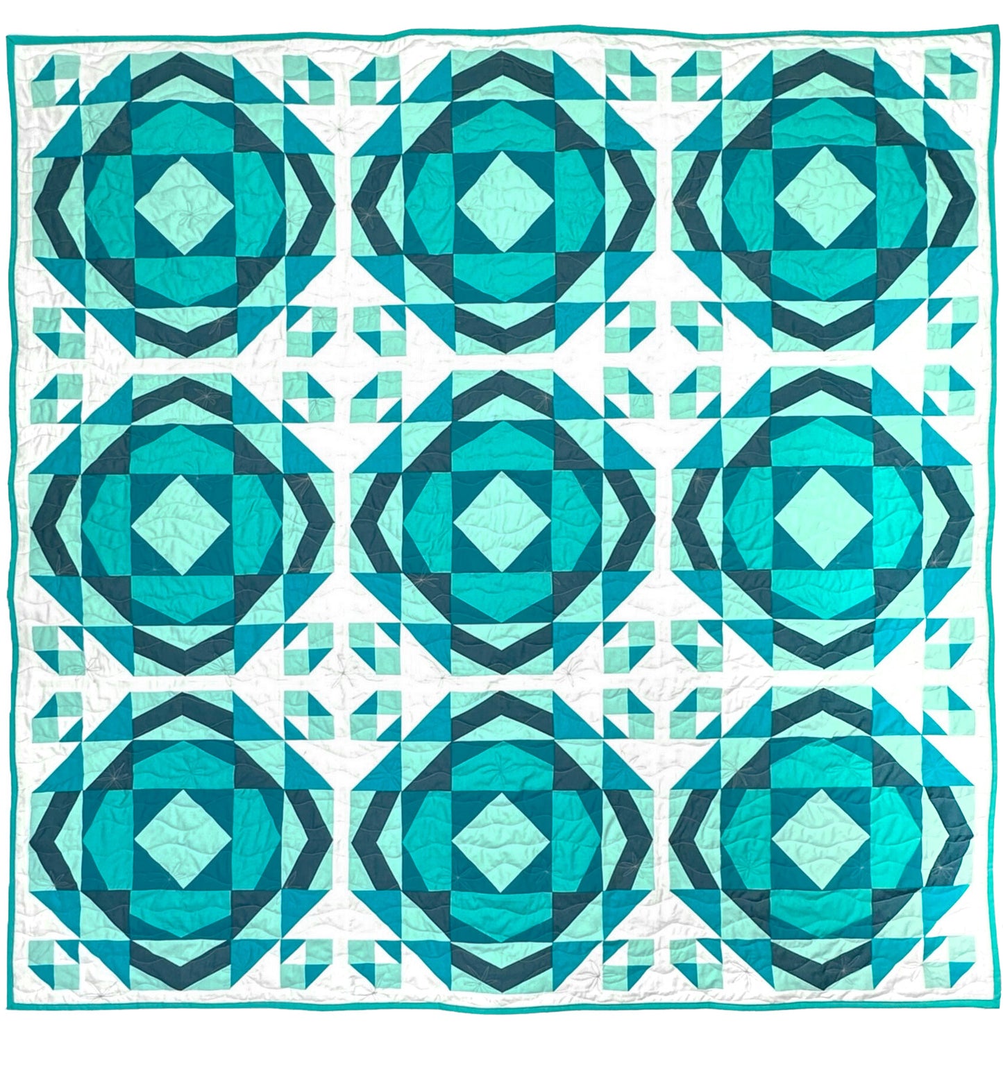 Millcreek quilt. A geometric quilt with a repeating motif that mimics water ripples. Made in varied shades of teal blue with a white background. Displayed flat on a white surface.