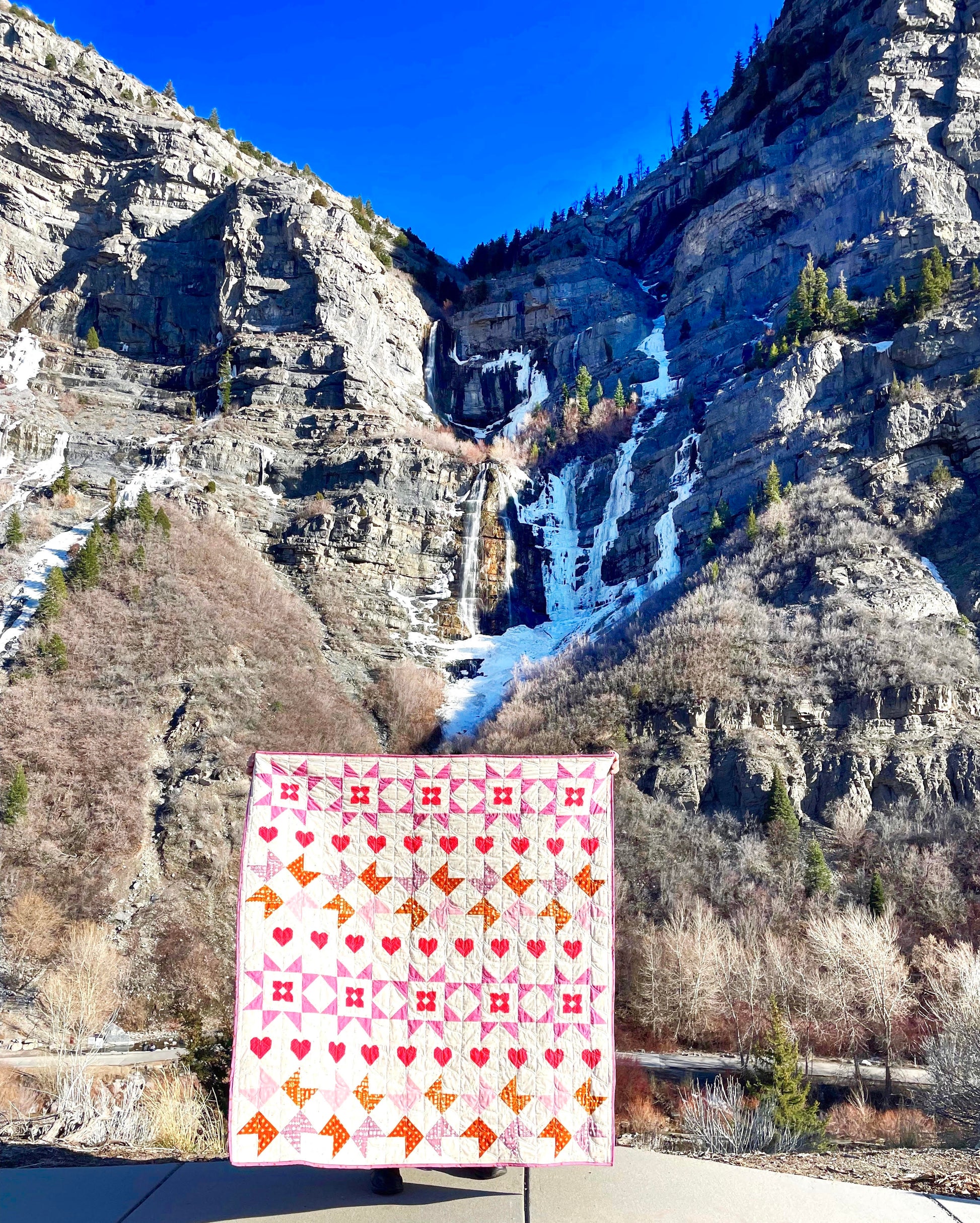 Nyla Star quilt. A star quilt with heart motifs and a flower within some star blocks. Made of pink, purple, and brown fabric on a light background. Displayed outdoors in front of a waterfall in a canyon.