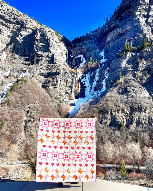 Nyla Star quilt. A star quilt with heart motifs and a flower within some star blocks. Made of pink, purple, and brown fabric on a light background. Displayed outdoors in front of a waterfall in a canyon.