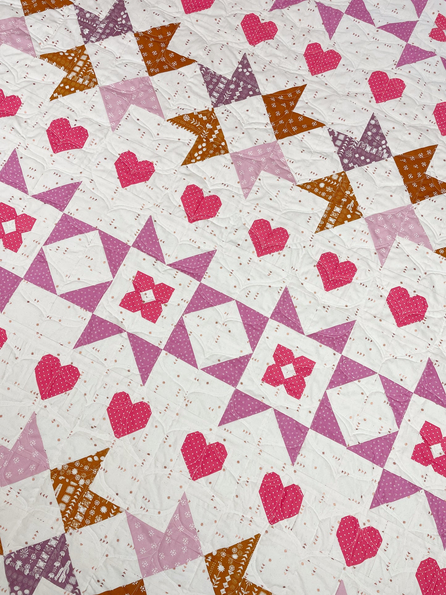 Nyla Star quilt. A star quilt with heart motifs and a flower within some star blocks. Made of pink, purple, and brown fabric on a light background. Displayed flat on an angle.