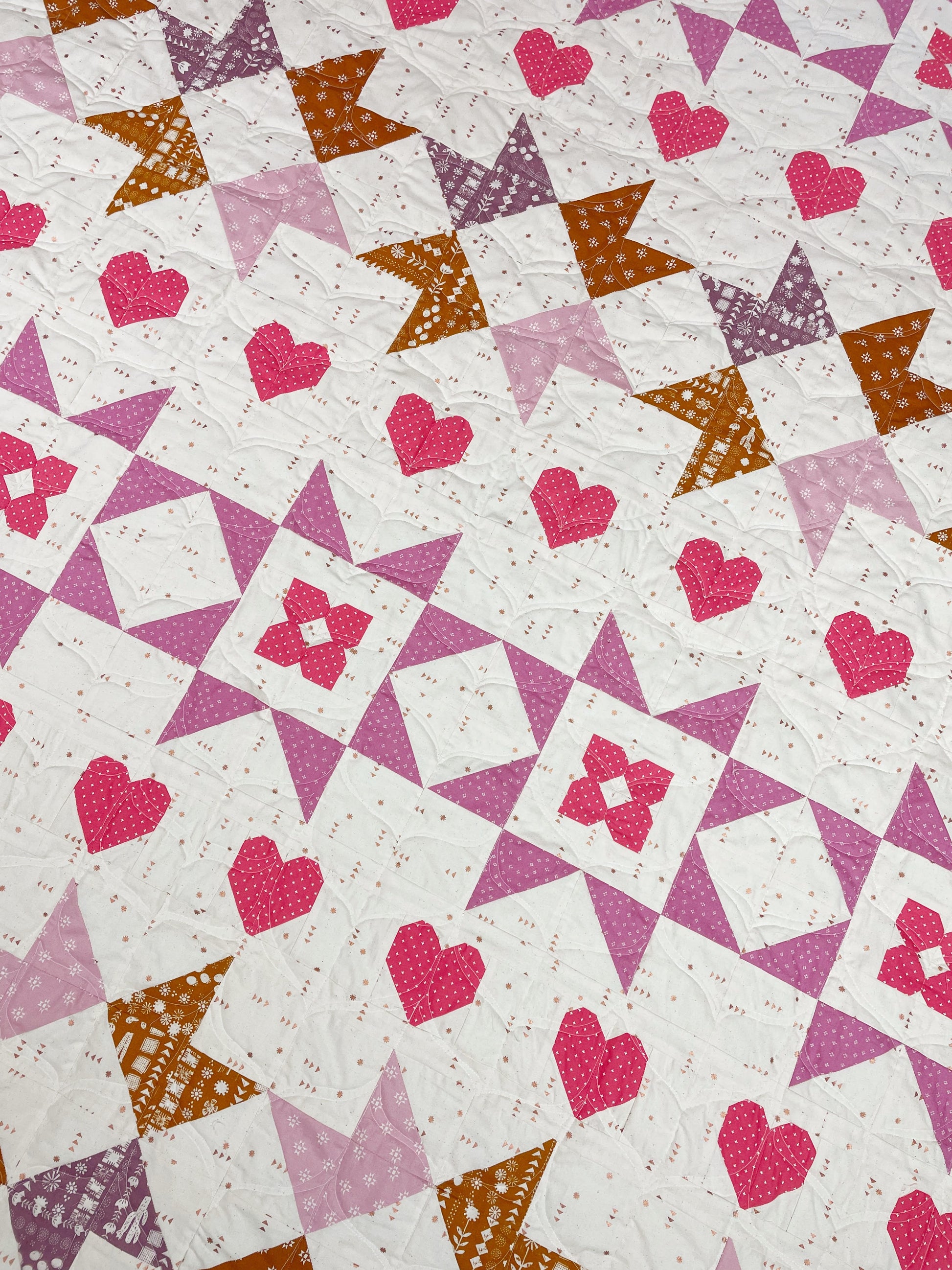 Nyla Star quilt. A star quilt with heart motifs and a flower within some star blocks. Made of pink, purple, and brown fabric on a light background. Displayed flat on an angle.