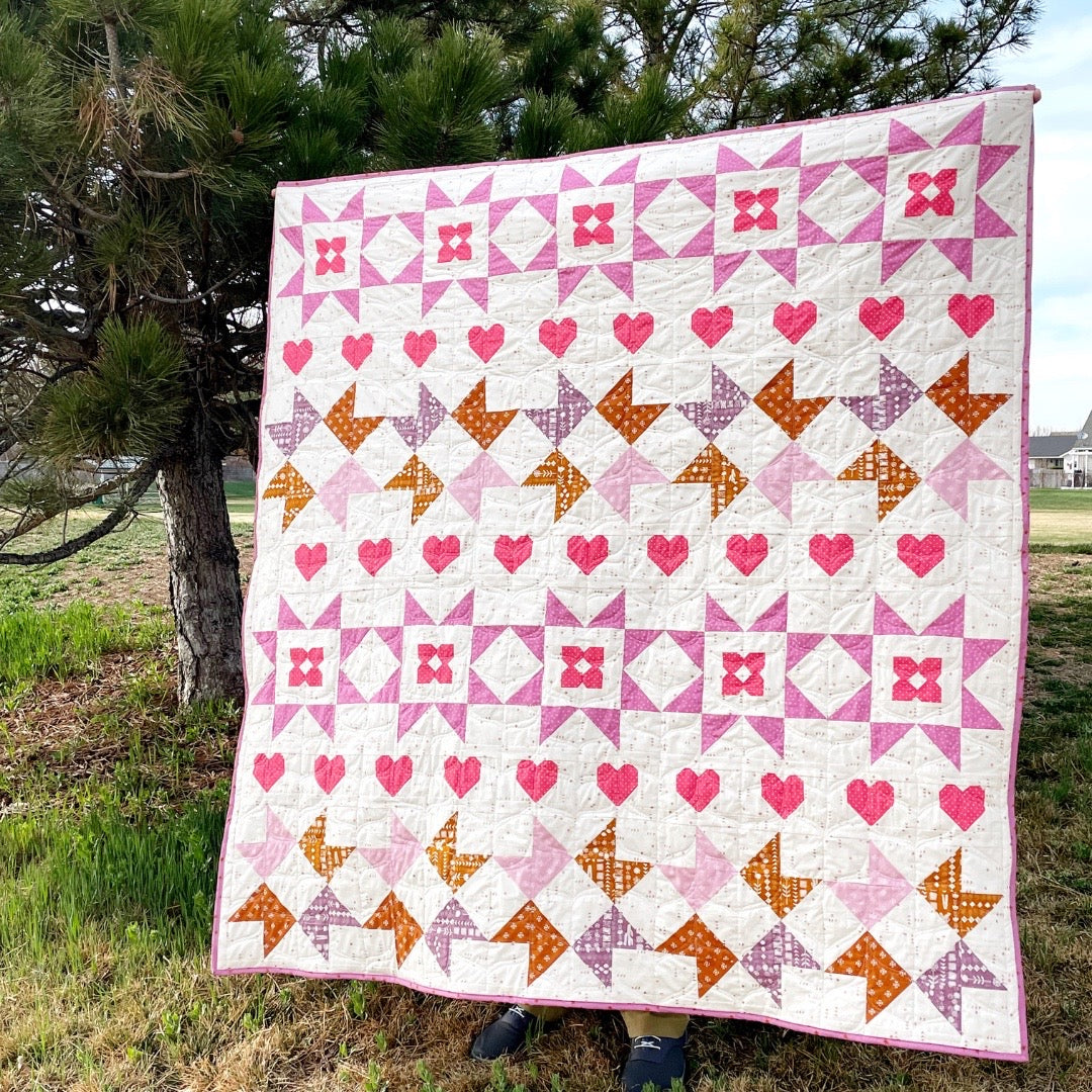 Nyla Star quilt. A star quilt with heart motifs and a flower within some star blocks. Made of pink, purple, and brown fabric on a light background. Displayed outdoors by a pine tree.