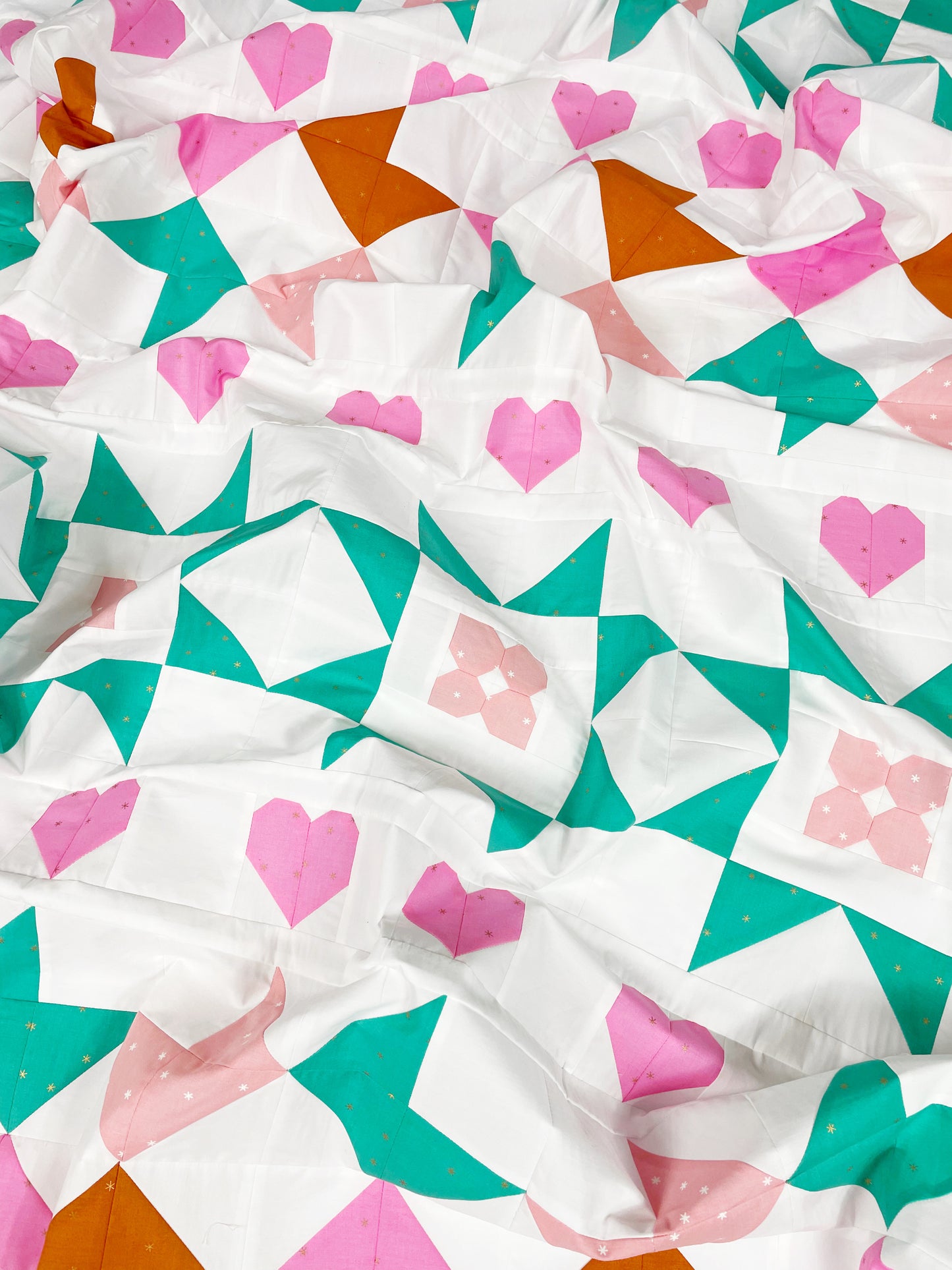 Nyla Star quilt. A star quilt with heart motifs and a flower within some star blocks. Made of pink, teal, and brown fabric on a light background. Displayed flat and slightly crumpled.