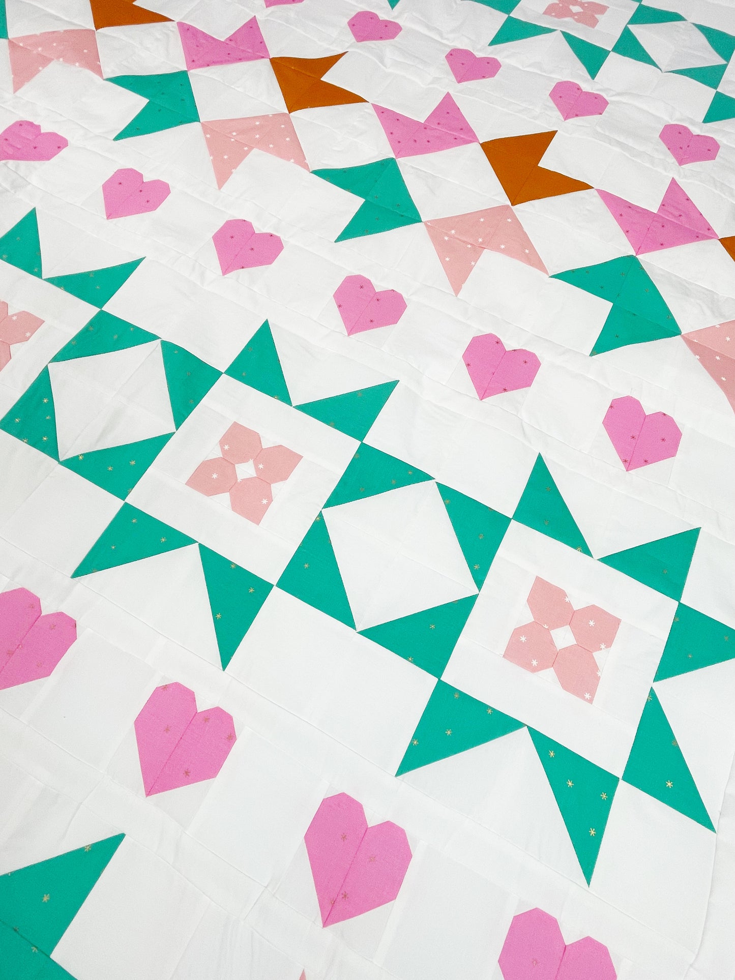Nyla Star quilt. A star quilt with heart motifs and a flower within some star blocks. Made of pink, teal, and brown fabric on a light background. Displayed flat and on an angle.