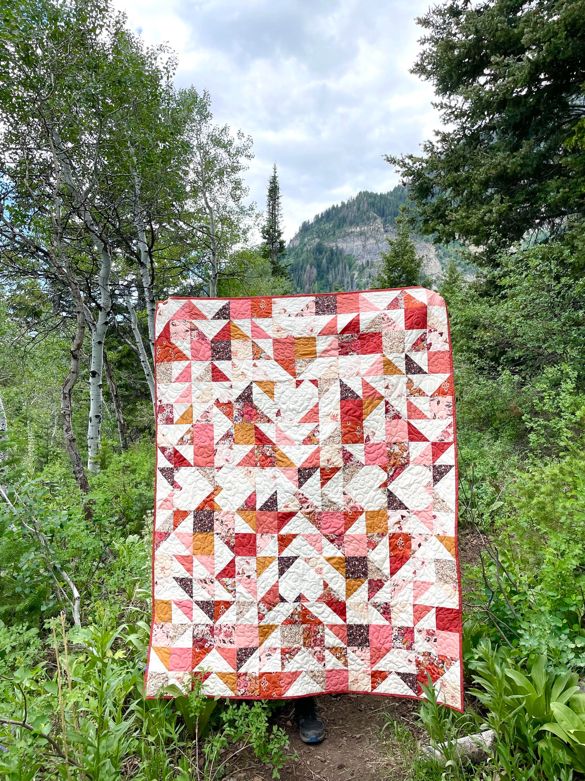Sweet LaNora quilt. A geometric quilt with triangle and heart motifs made of fabric with red and peach tones.  Displayed outdoor in a mountainous landscape with trees.