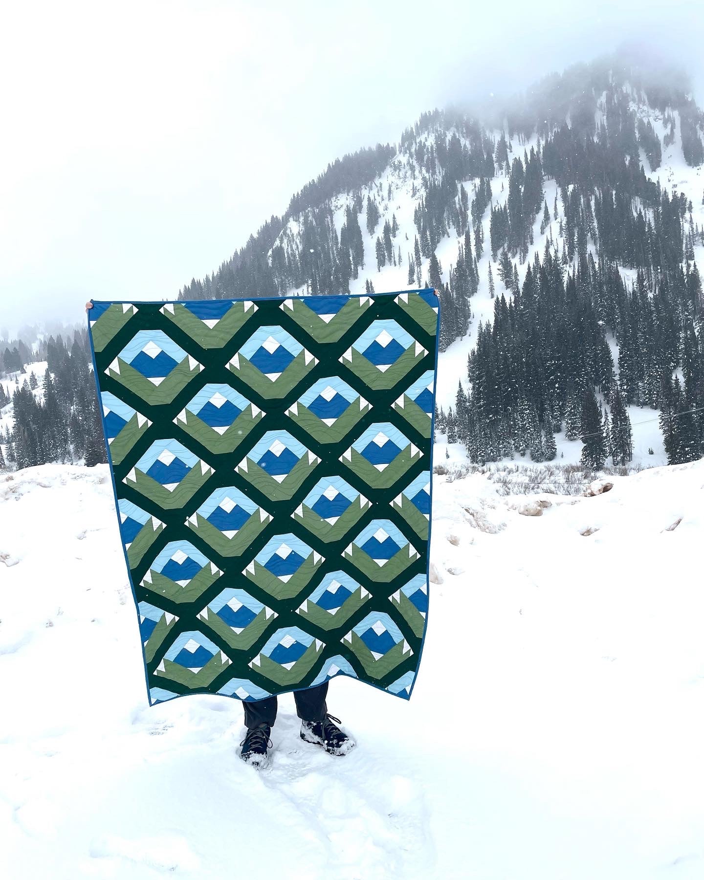 Wasatch quilt. A quilt with a repeating motif that appears similar to a mountain capped with snow above green strips. Blocks set on point. Made with green, blue, and white fabric. Displayed outdoors in a snowy, mountainous landscape.