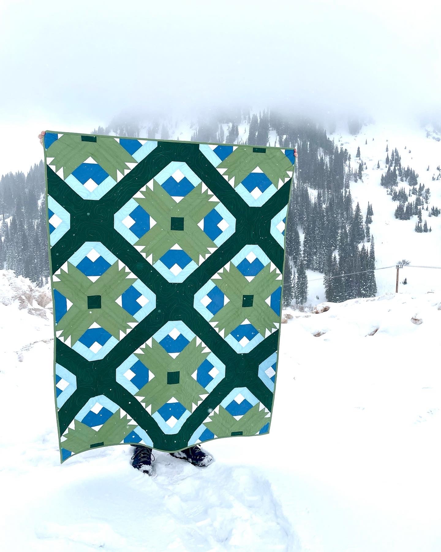 Wasatch quilt. A quilt with a repeating motif that appears similar to a mountain capped with snow above green strips. Blocks set on point. Rotated so blocks appear as a trellis design. Made with green, blue, and white fabric. Displayed outdoors in a snowy, mountainous landscape.